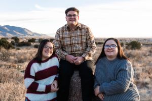 Three people wearing sweaters pose together in a desert landscape with mountains in the background.