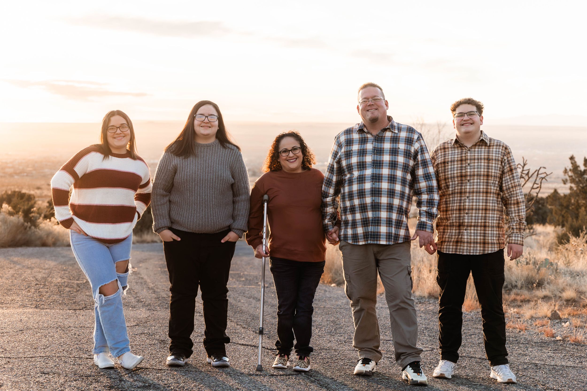 A family group stands together smiling on a dirt path at sunset with desert brush in the background.
