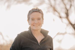 A person wearing a black zip-up jacket smiles warmly against a soft, sun-lit natural background.