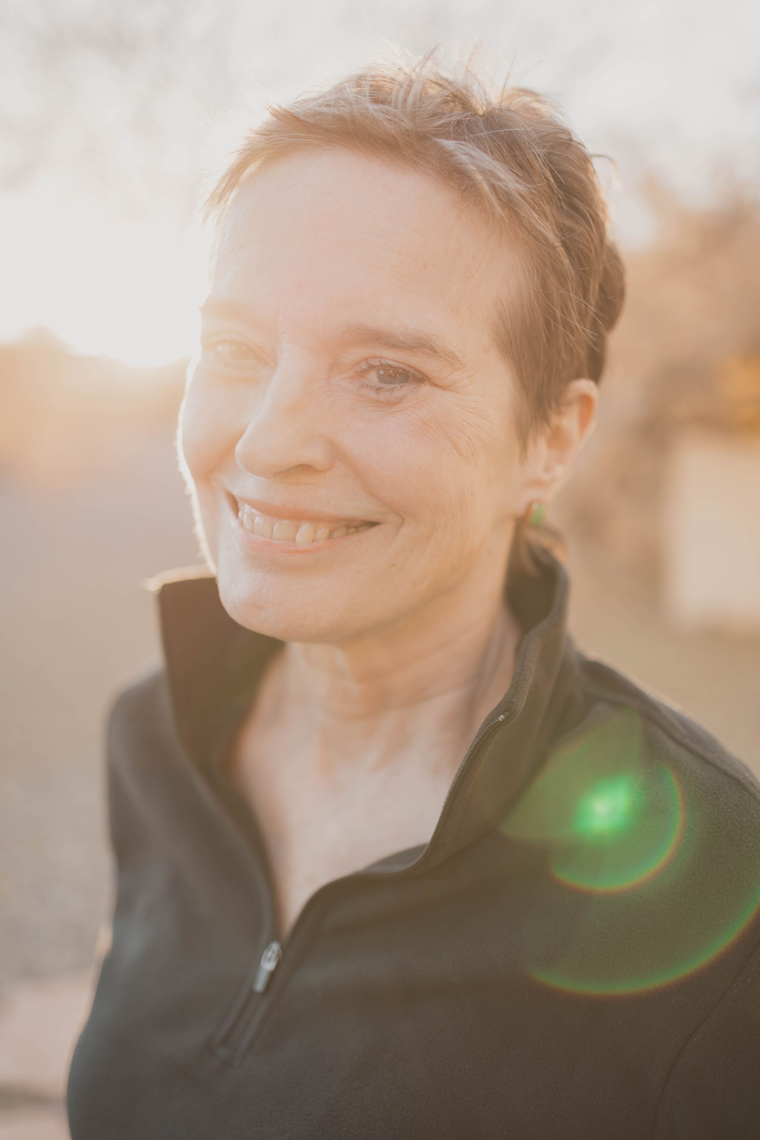 A person smiles warmly while wearing a black zip-up jacket in soft sunlit lighting with lens flare.