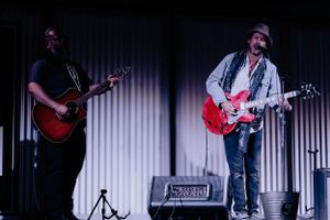 Musicians, Nate Fox and Sale, perform on stage with acoustic and electric guitars in front of an amplifier and dark curtain backdrop.