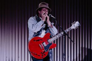 Musician< Nathan Fox,  performs on stage with a red electric guitar while wearing a gray hoodie and brown hat.