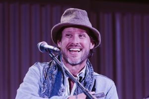 Performer, Nathan Fox Band, in a fedora hat and patterned vest smiles while standing at a microphone on stage.