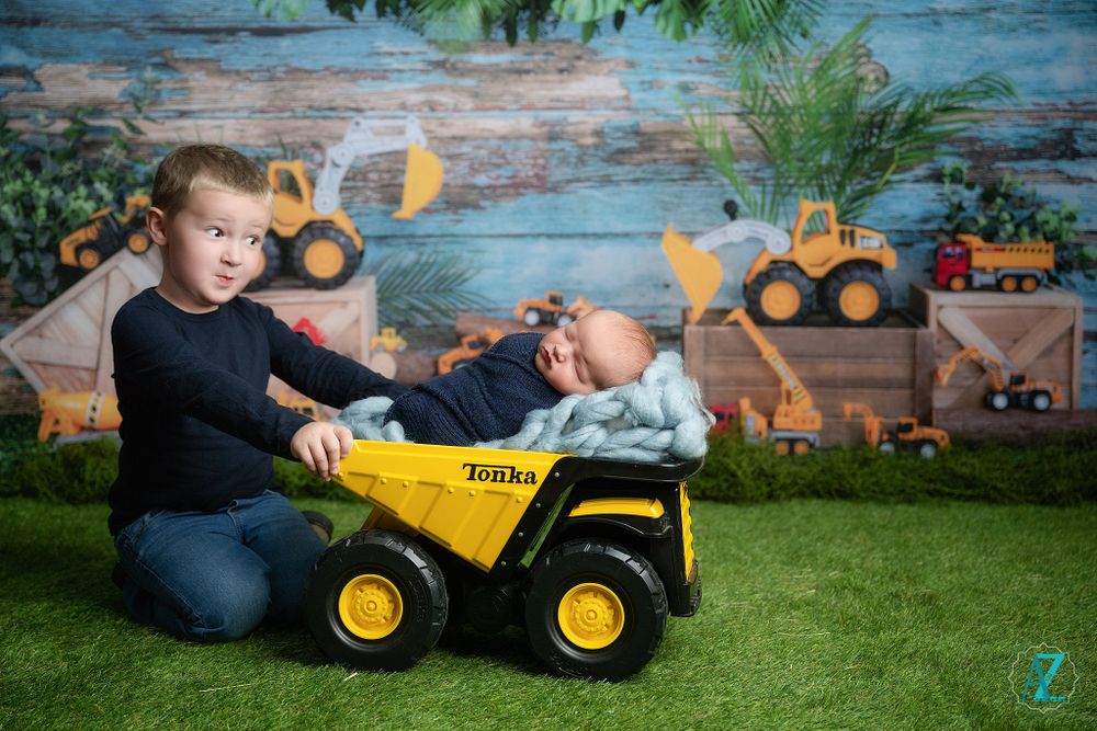 Children playing with a yellow toy dump truck against a rustic backdrop with construction vehicle decorations.