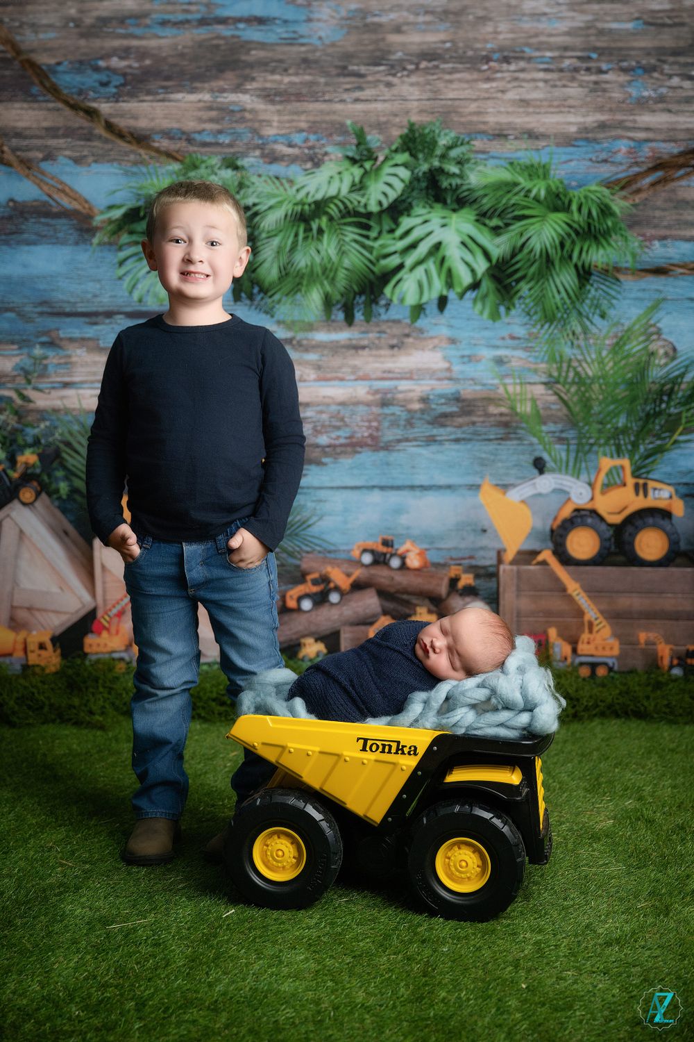 Child stands proudly next to yellow toy truck against construction-themed backdrop with greenery.