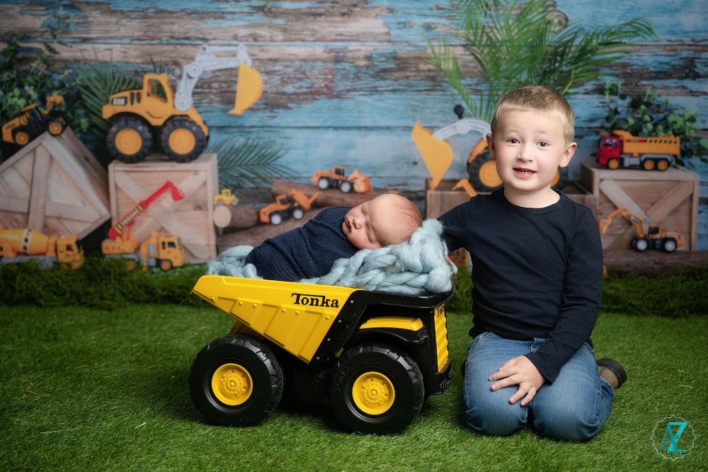 Two children pose with a yellow Tonka dump truck toy against a decorated construction-themed backdrop.