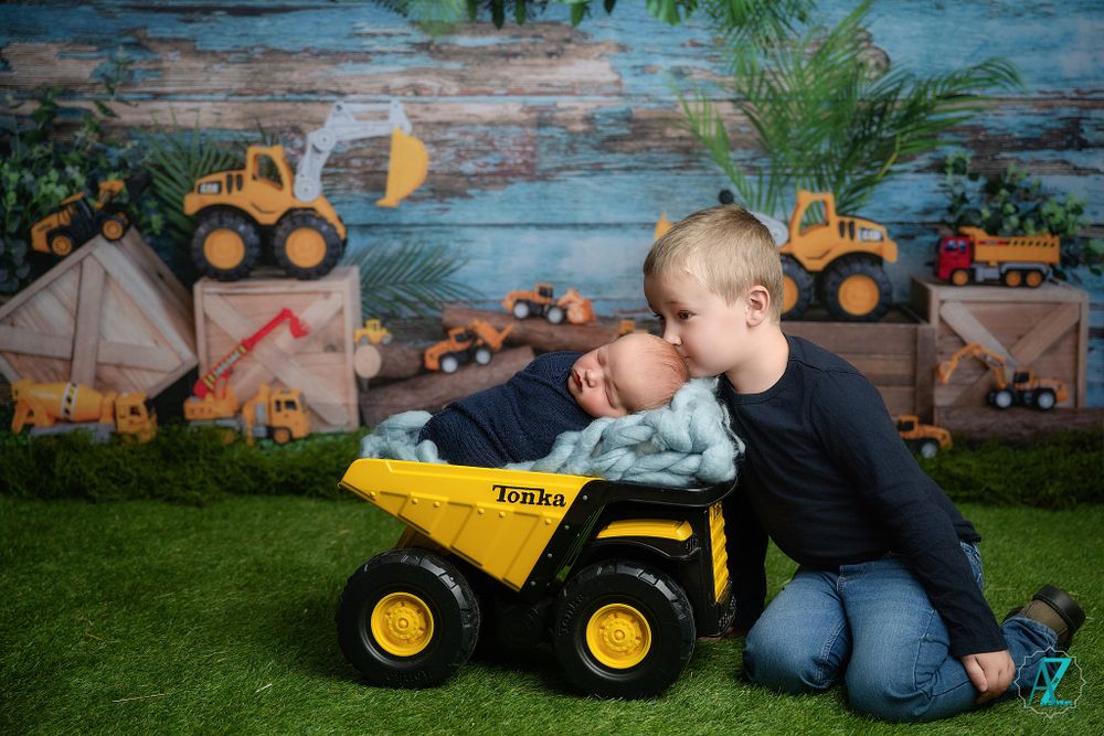 Child plays with yellow toy dump truck on artificial grass with construction-themed backdrop.