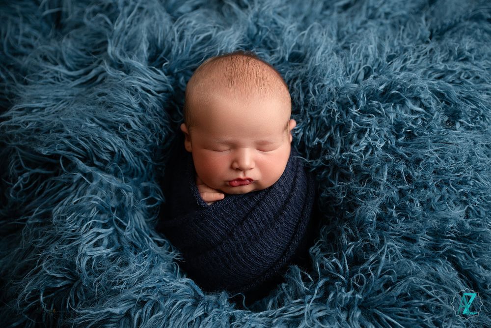 Sleeping newborn wrapped in navy knit wrap on blue fluffy textured backdrop.