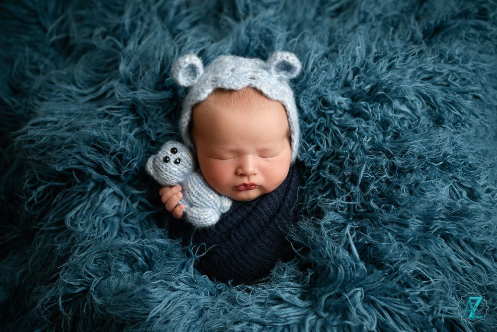 Sleeping newborn wearing bear ear headband with stuffed toy on blue furry backdrop.