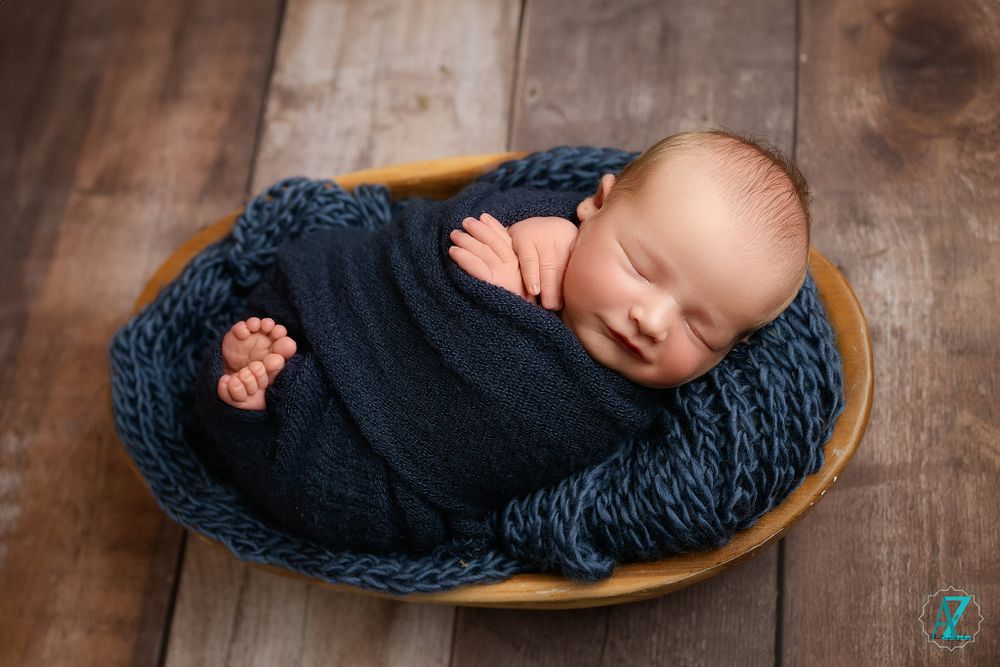 Sleeping newborn wrapped in navy blanket lying in wooden bowl with textured backdrop.