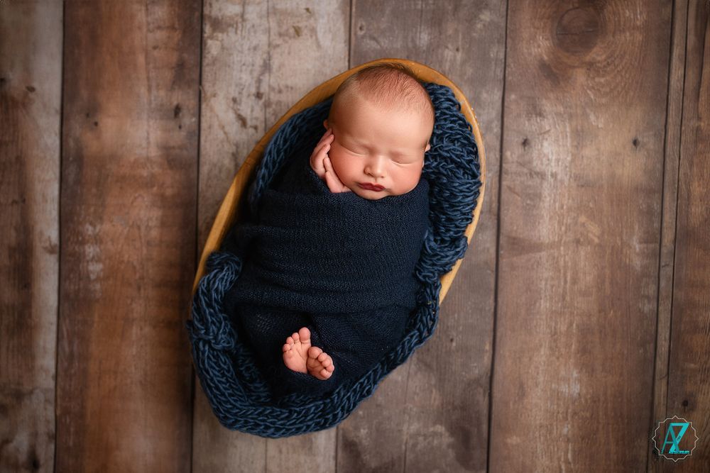 Peaceful sleeping newborn wrapped in navy knit blanket in wooden bowl against rustic background.