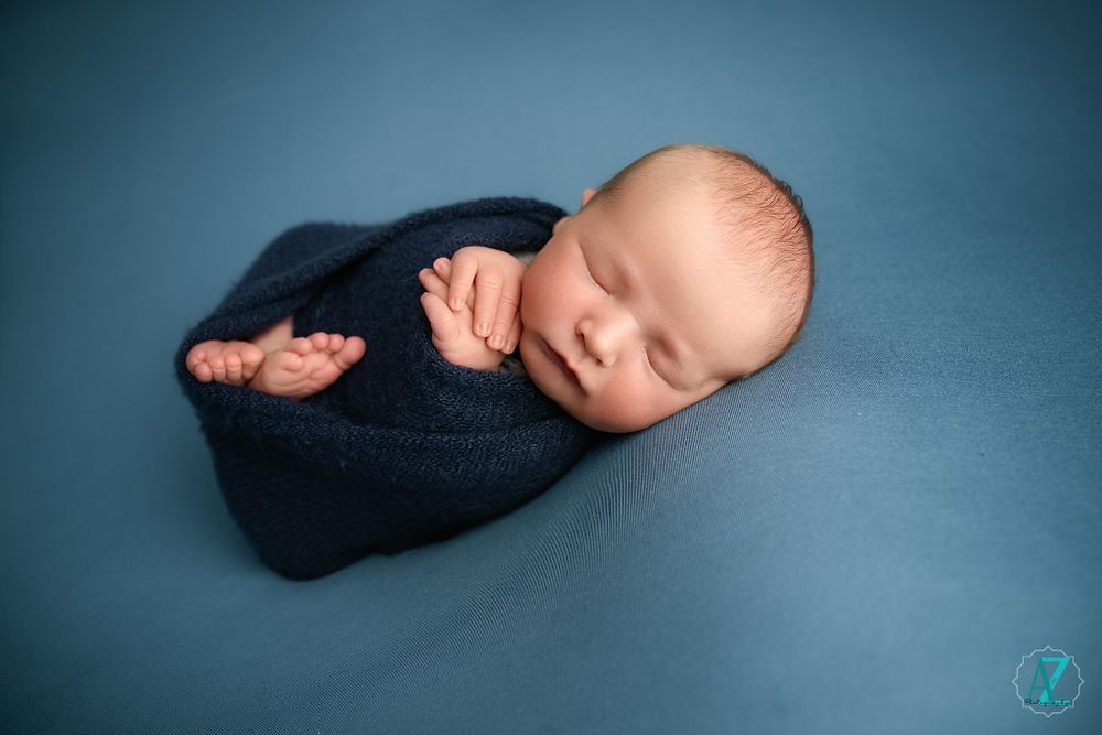 Peaceful newborn swaddled in navy blue blanket sleeping against blue backdrop during professional photo session.