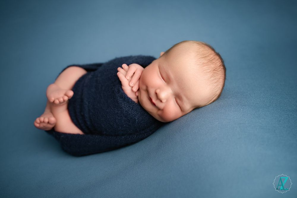 Sleeping baby wrapped in dark blue swaddle on blue background during newborn photoshoot.