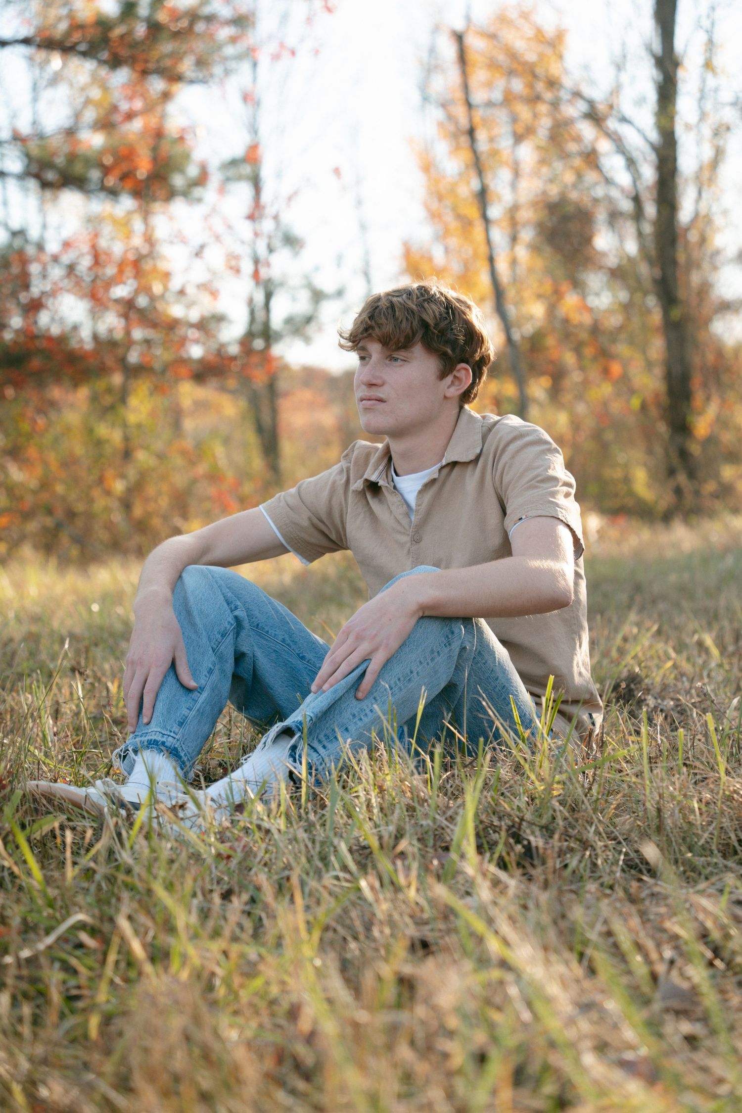 Young man in beige shirt and jeans sitting on grass in autumn park with colorful fall foliage background.