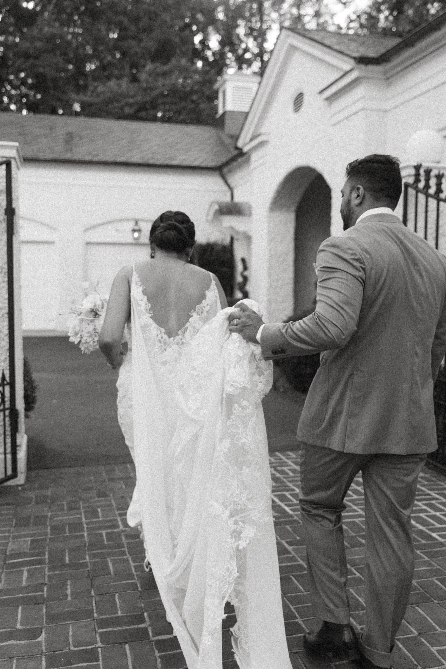 Couple walking through arched entrance of white building with bride's lace dress trailing behind.