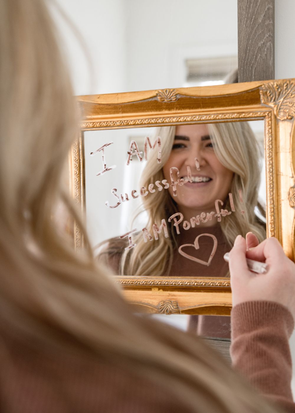 Person writing on mirror with lipstick in ornate gold frame.