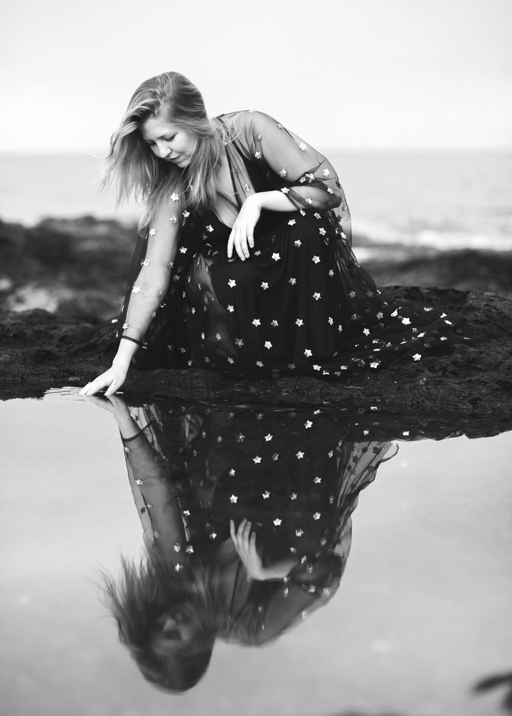 Black and white reflection photo with person in polka dot dress on rocky shore.