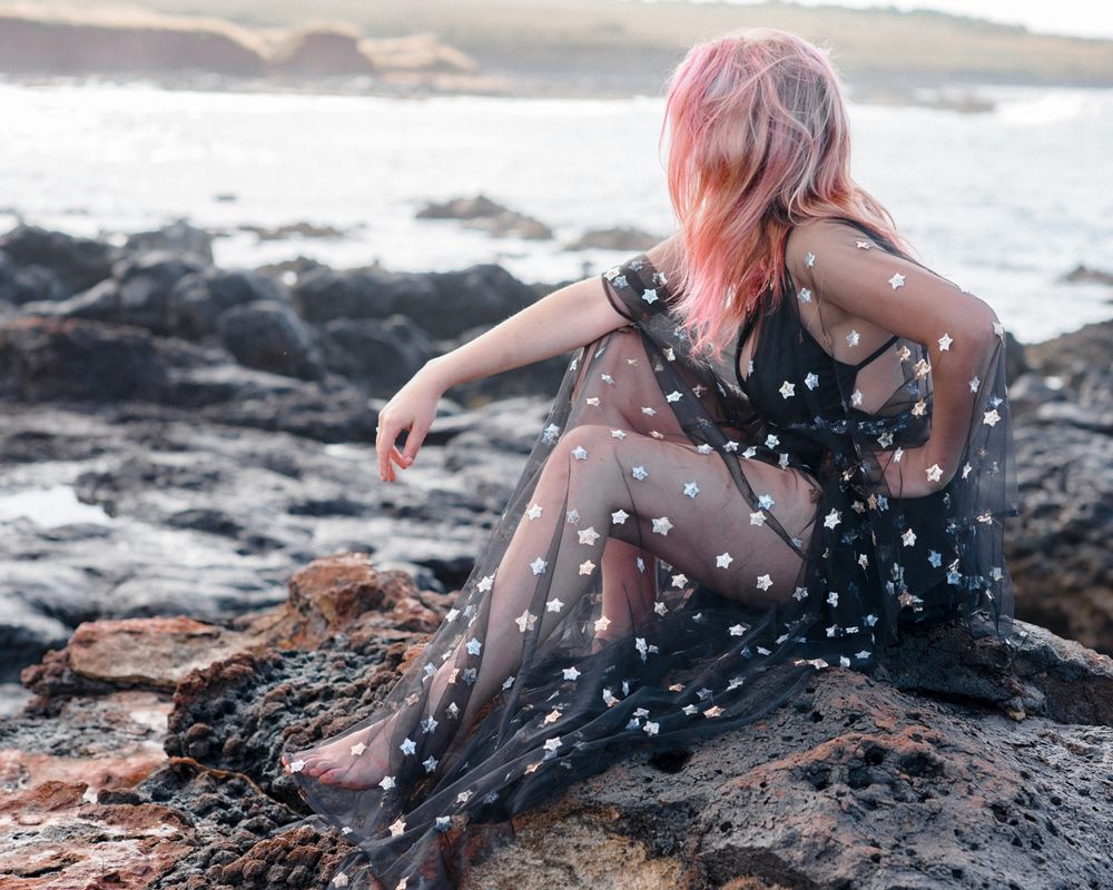 Person with pink hair in sheer polka dot dress poses on rocky coastline.
