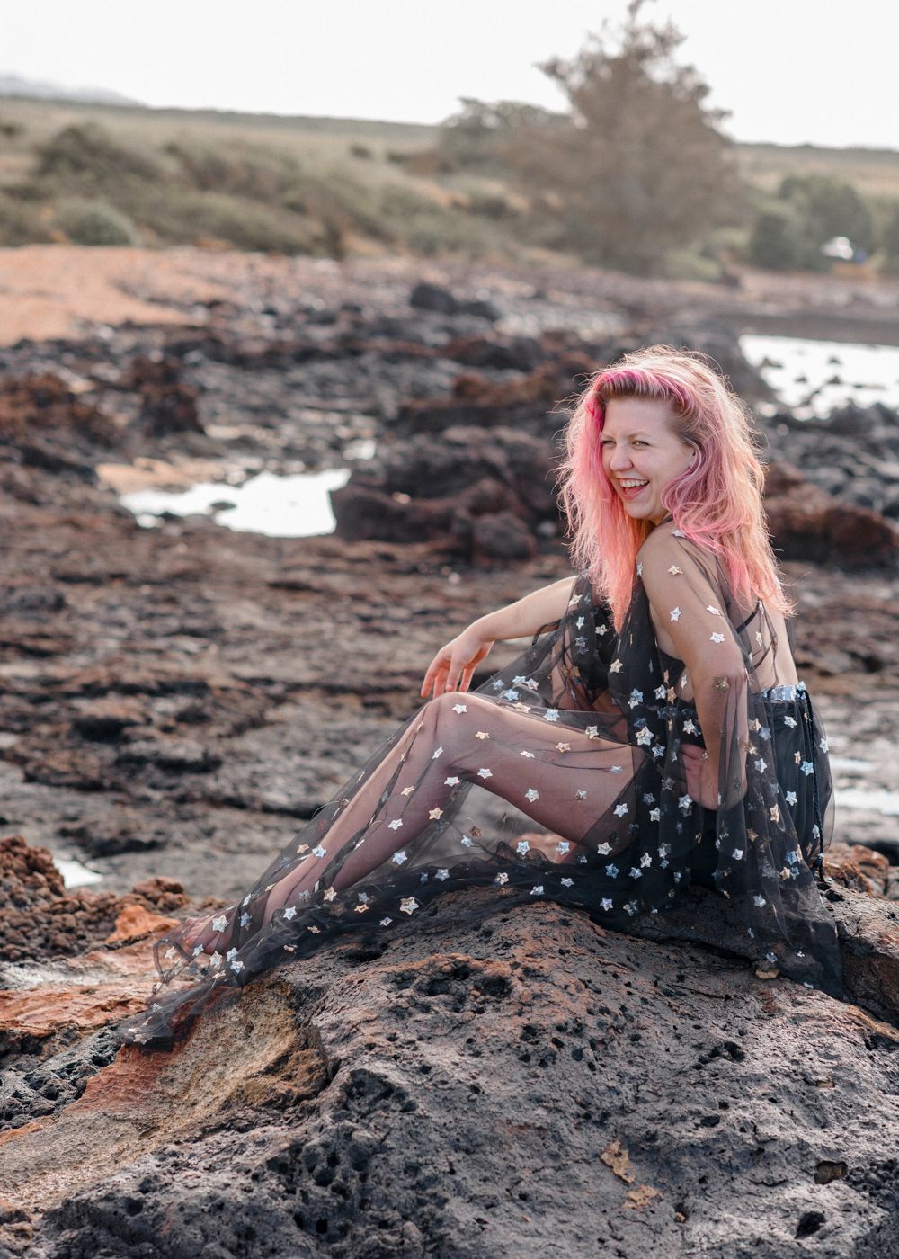 Pink-haired model in polka dot dress sitting on volcanic rocks by ocean.