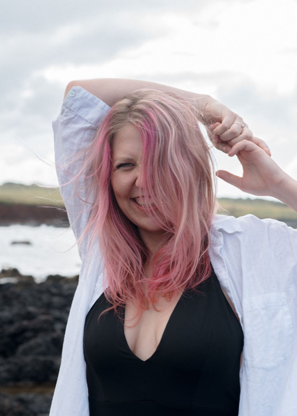 Person with pink hair and white shirt poses against rocky coastal backdrop.