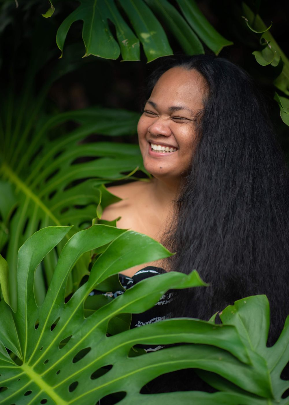 Person with long dark hair smiling among tropical monstera leaves.