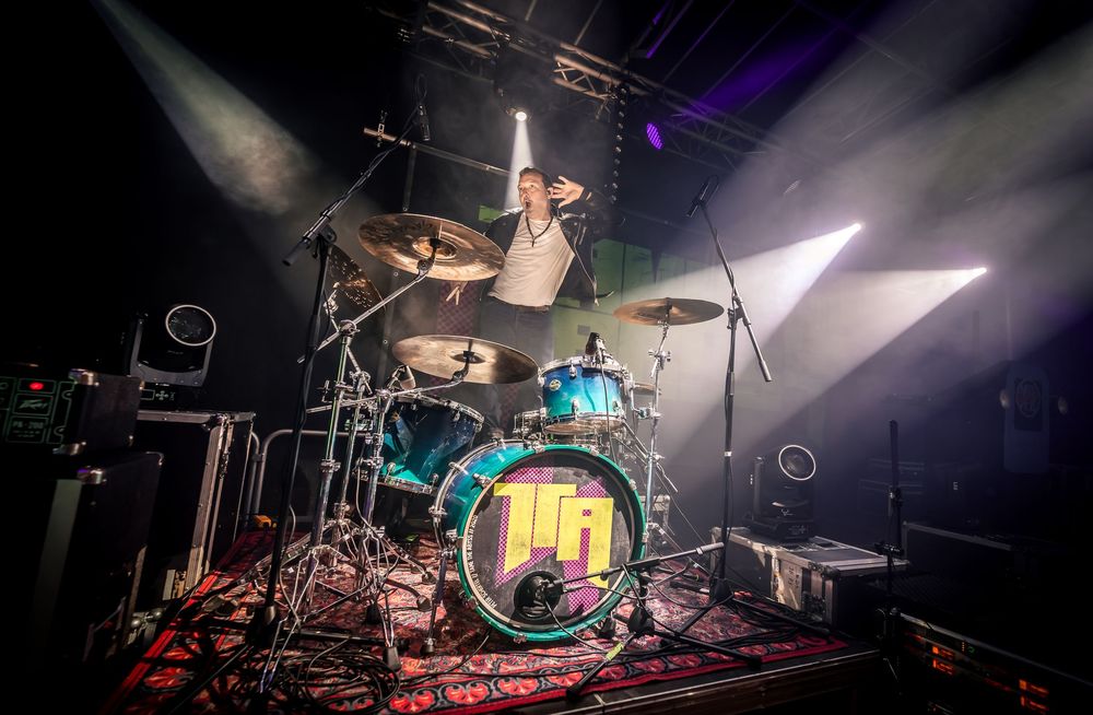 A panoramic view of a concert stage setup with drums, lighting equipment, and musical instruments in a dark venue.