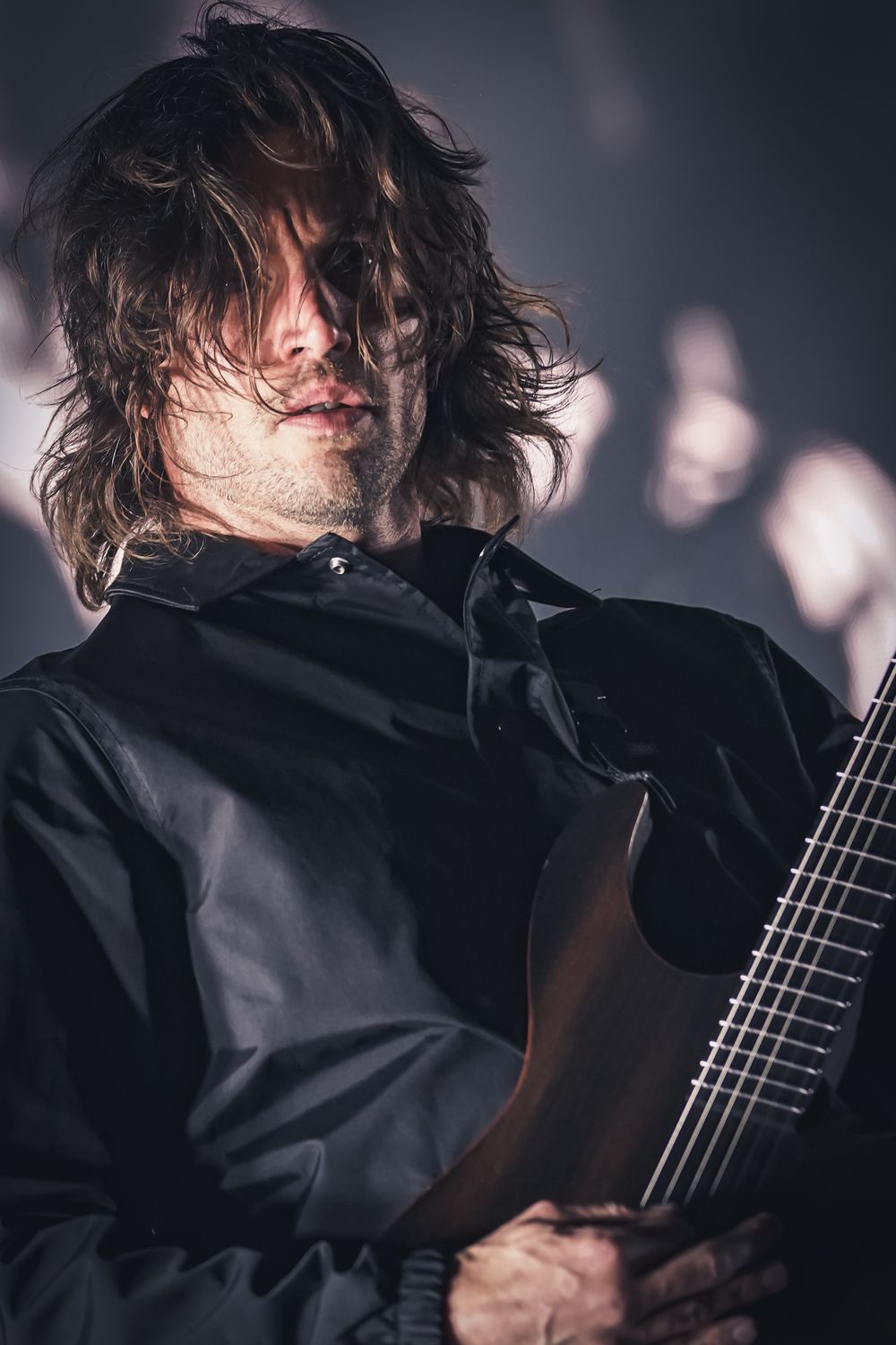 Musician in black clothing performs on stage with guitar in dramatic lighting with long tousled hair.