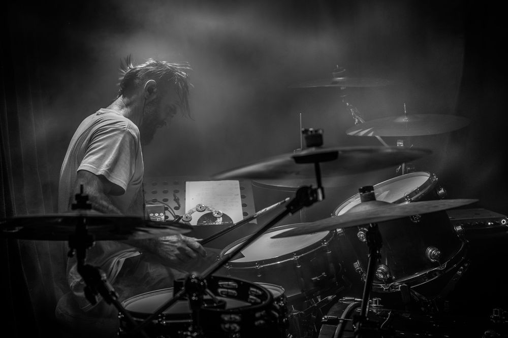 A drummer performs on stage in dramatic black and white lighting during a live music performance.