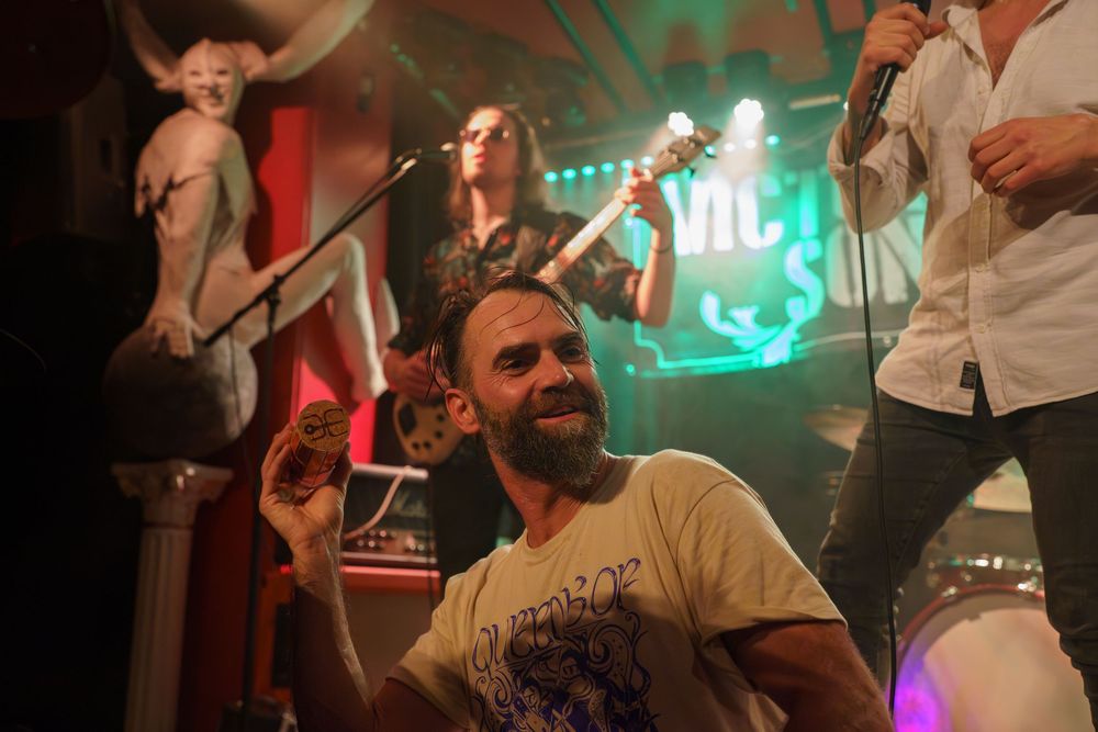 A performer with a beard in a white t-shirt smiles on stage while musicians play in a dimly lit venue.
