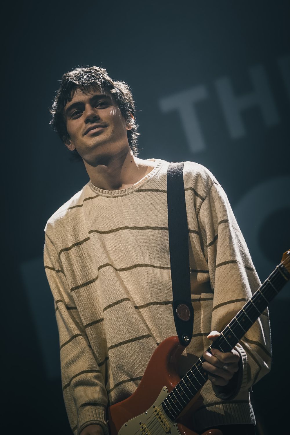A guitarist in a beige sweater performs on stage under dramatic lighting while wearing sunglasses.