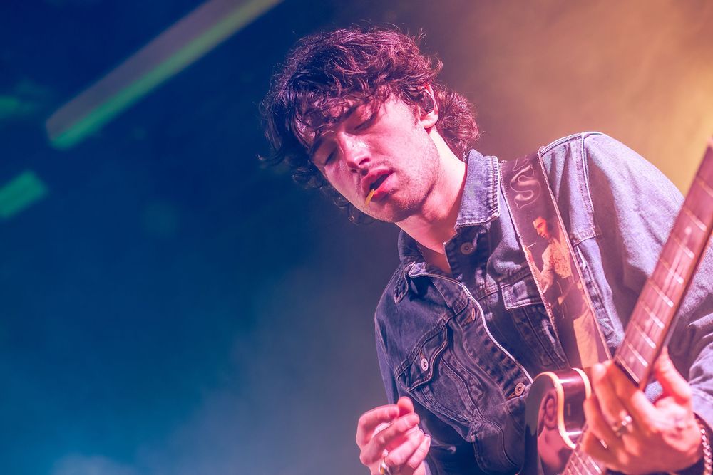 A guitarist performs on stage with dramatic blue lighting and energetic movement while playing an electric guitar.