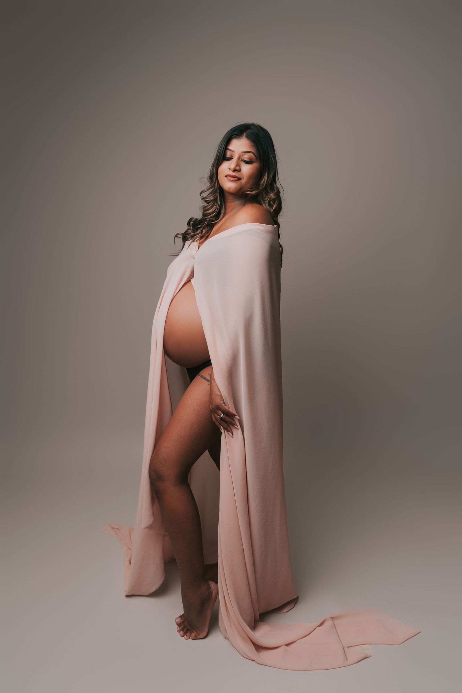 Artistic maternity photos in blush tones showing a pregnant figure draped in flowing fabric against neutral backdrop.