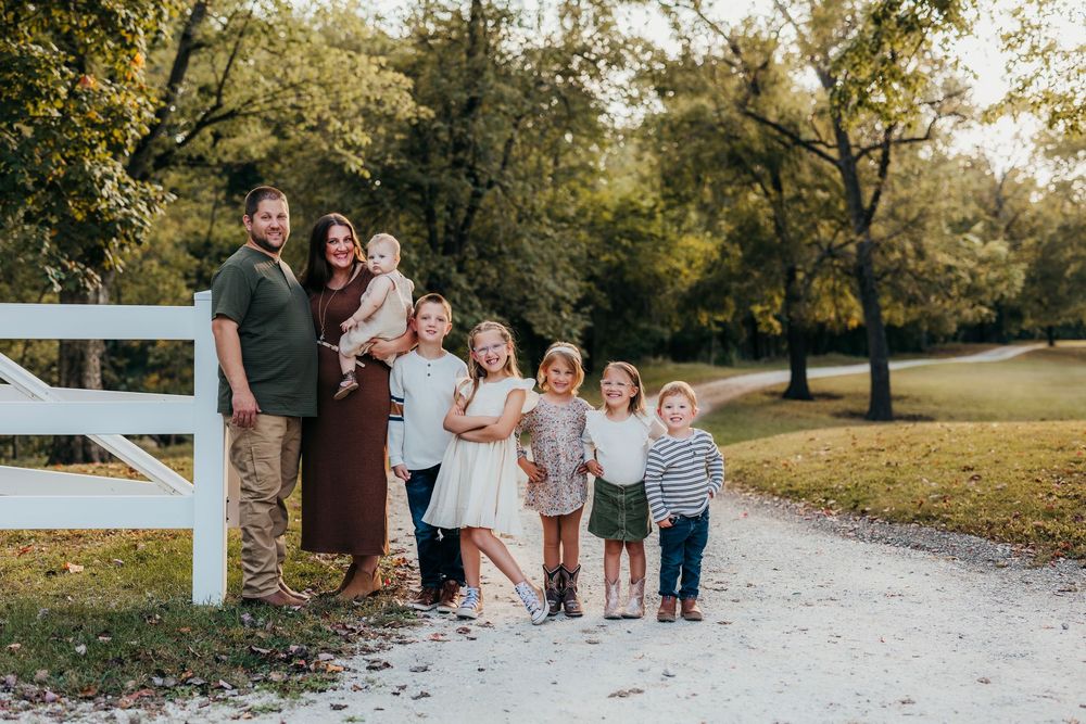 A group gathers for outdoor autumn family portraits along a white fence and gravel path in golden sunlight.