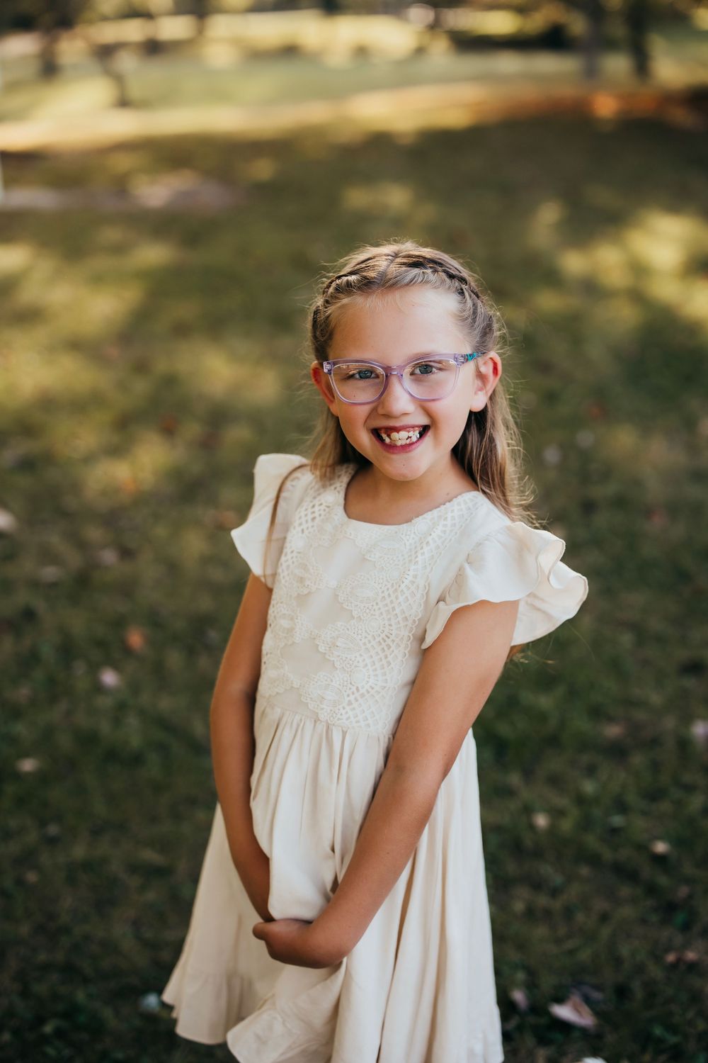 Young child in a white lace dress smiling brightly while wearing glasses in an outdoor garden setting.