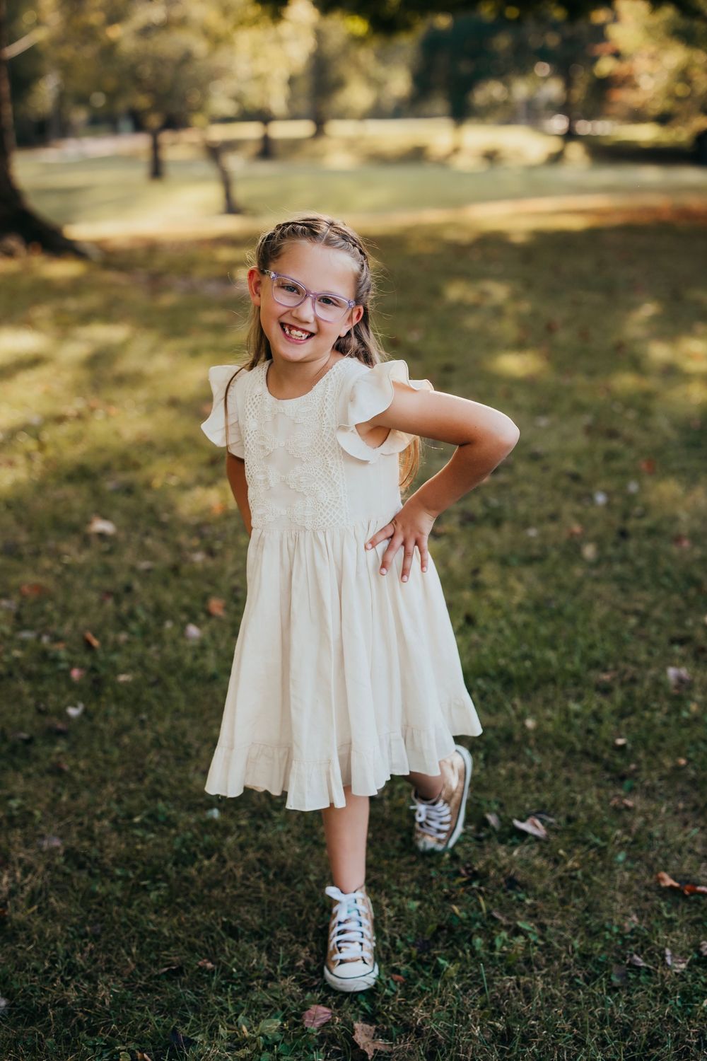 Young child in white dress sitting playfully on grass during outdoor photo session.