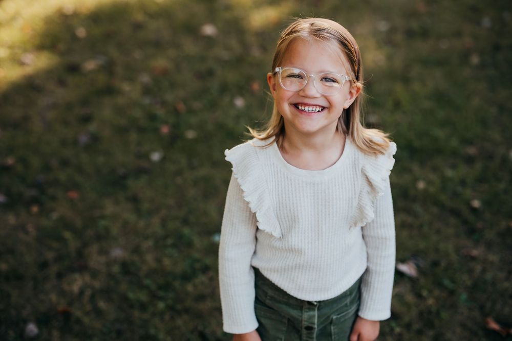 Child wearing glasses and a white dress poses joyfully against a natural outdoor background.