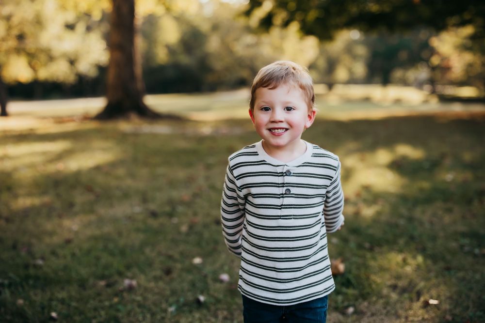 A child in a gray striped shirt laughs joyfully while playing outdoors on a sunny autumn day.