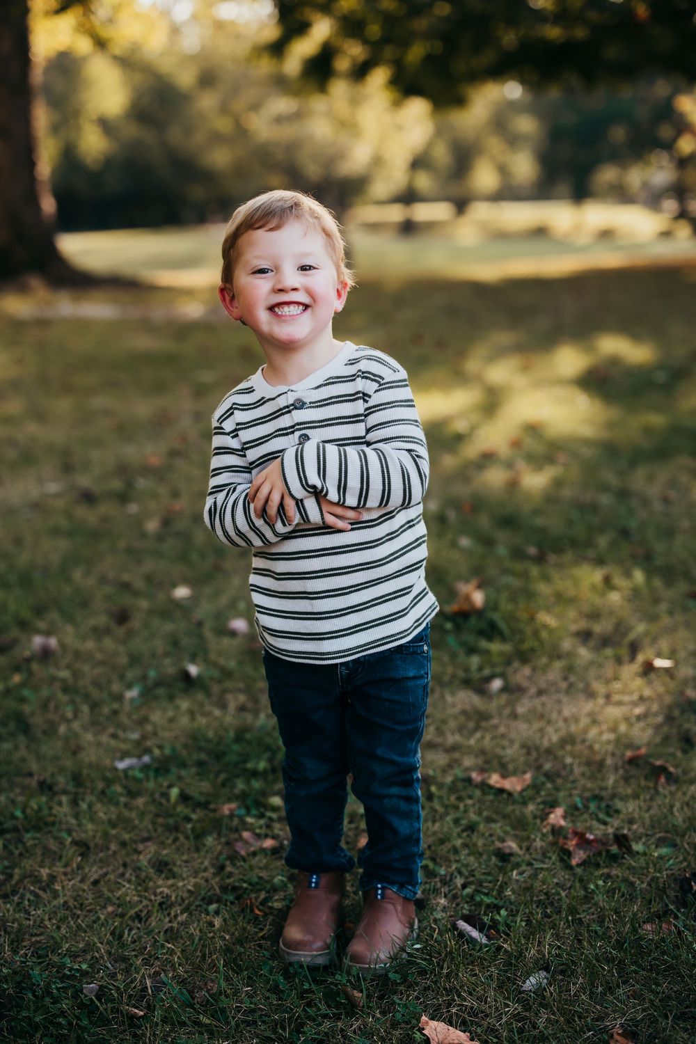 A toddler in a striped shirt and jeans plays outdoors on grass during a sunny autumn day.