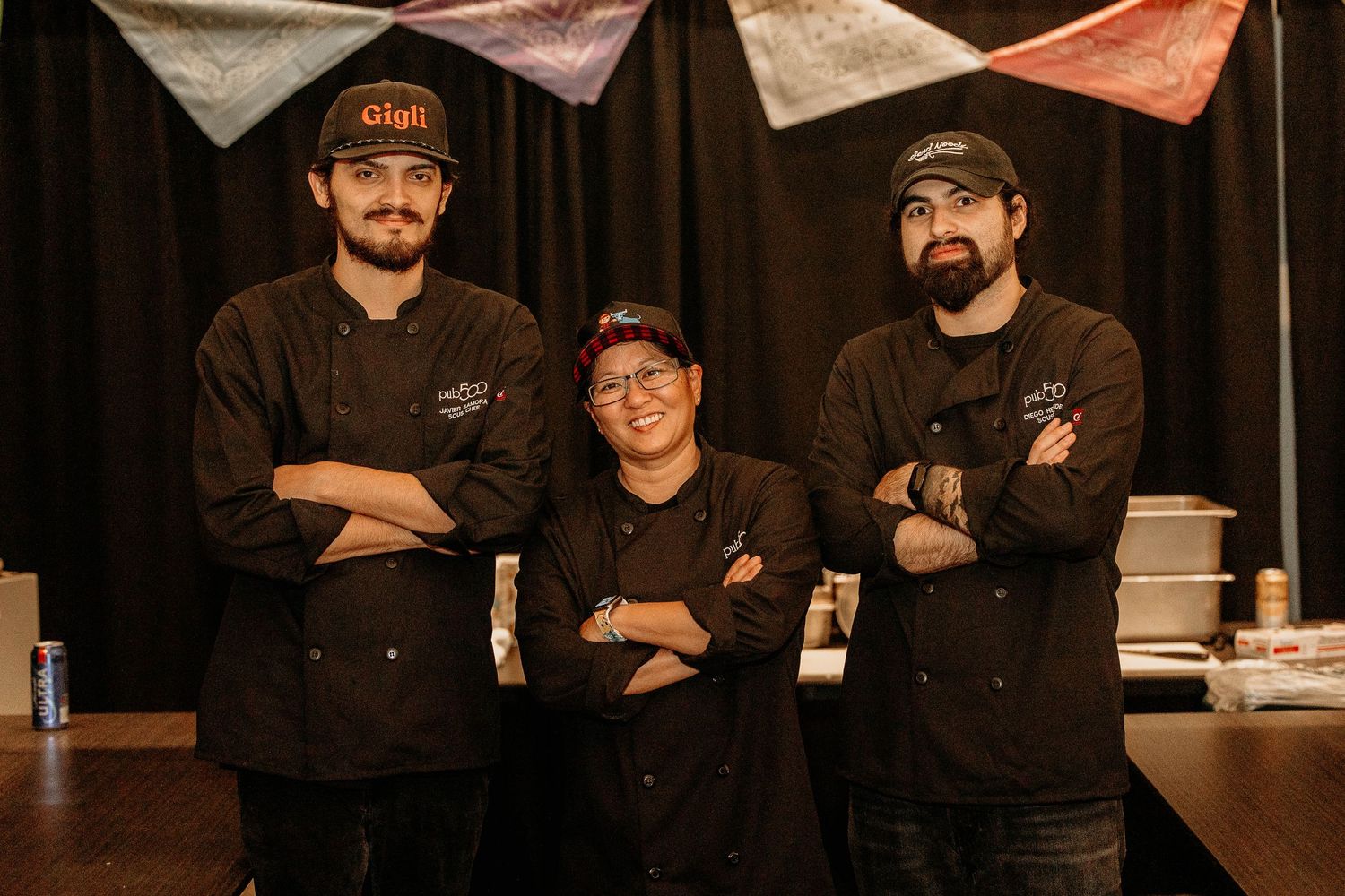 Three chefs in black uniforms stand together with crossed arms in front of a dark curtain backdrop.