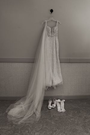 Wedding dress hanging with train and accessories arranged below on concrete floor.