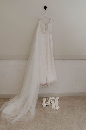 A white wedding dress hangs elegantly against a light wall with white gloves and shoes displayed below.