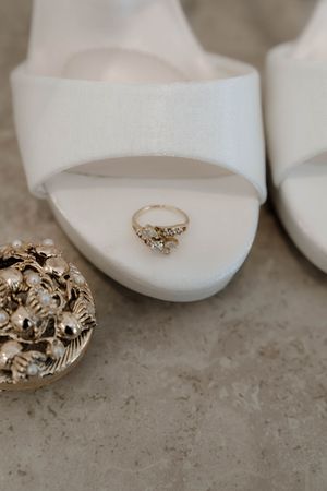 Ornate gold wedding rings displayed next to white bridal shoes on a textured surface.