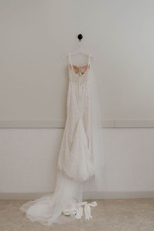 An elegant white wedding gown with train displayed on a hanger against a light wall.