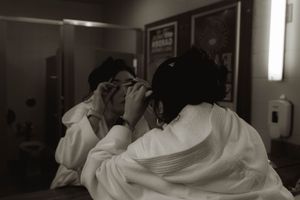 Black and white photo showing someone applying makeup in a dressing room mirror.
