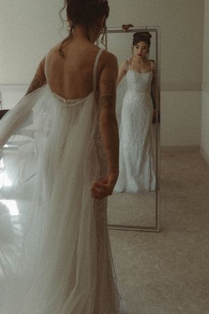 Bride in a flowing white gown looks at her reflection in a full-length mirror.