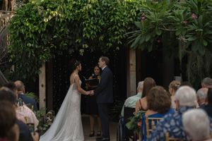 An outdoor wedding ceremony under lush green trees with guests seated on white chairs.