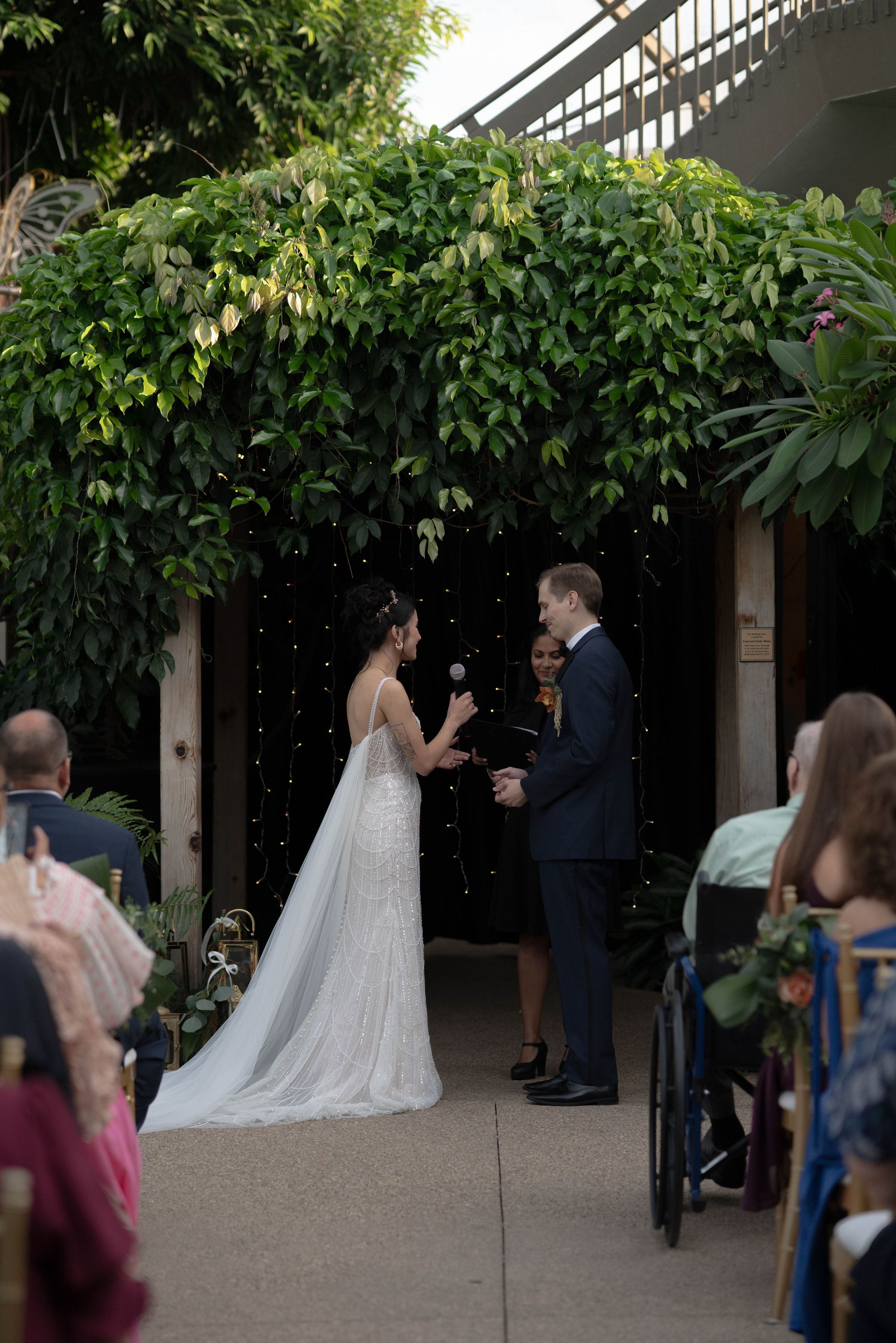 Outdoor wedding ceremony under a vine-covered pergola with guests seated on white chairs.