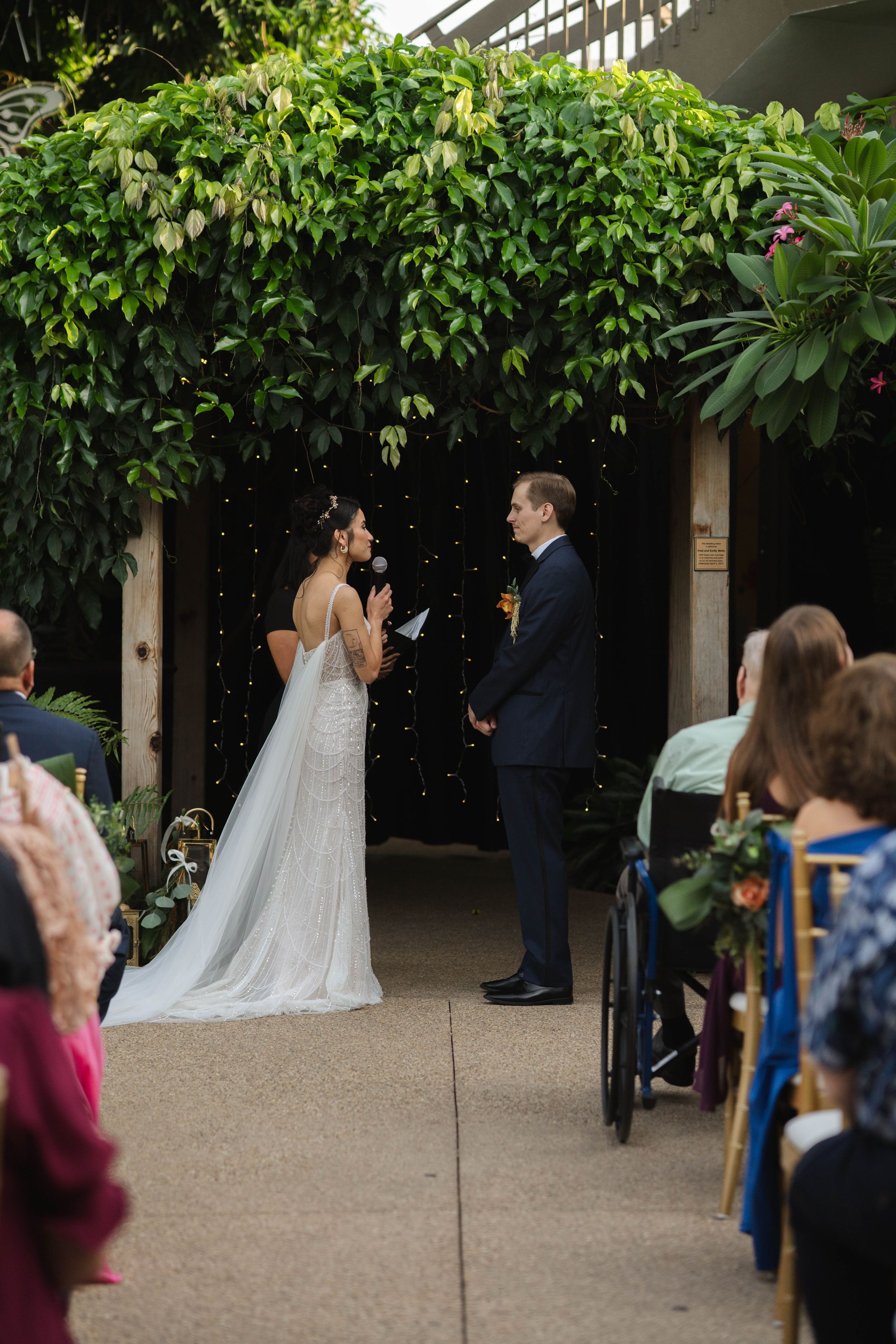 Wedding ceremony in progress under a leafy green pergola with guests watching.