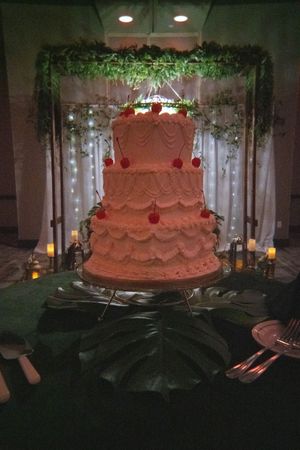 Elegant pink tiered wedding cake displayed under greenery arch with fairy lights and candles.
