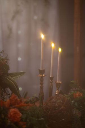 Three lit taper candles in brass holders surrounded by floral arrangements and soft bokeh.